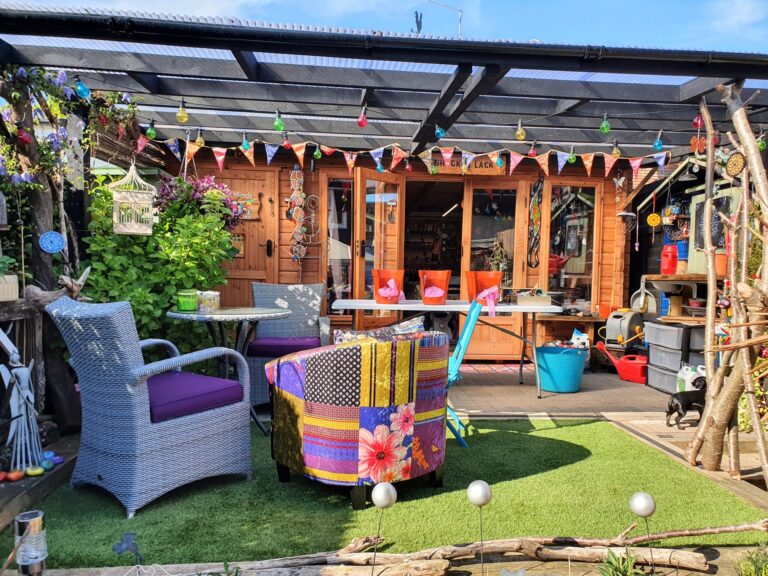 A colorful backyard garden setup featuring a wooden garden shed with large glass doors and festive bunting strung across the top. The patio area includes a variety of outdoor furniture: a wicker chair with a purple cushion, a bright patchwork armchair, and a white table surrounded by orange chairs. The space is decorated with hanging lanterns, potted plants, garden tools, and string lights. Artificial grass covers the ground, and a black-framed transparent roof provides partial shade. The area has a playful, cozy, and eclectic vibe.