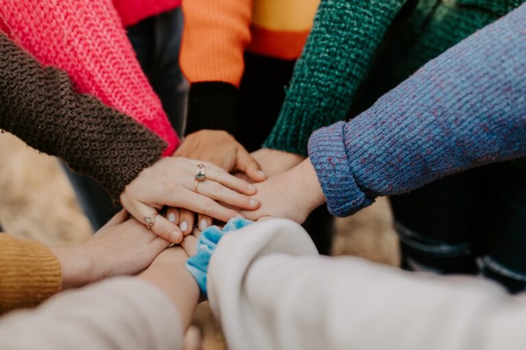 Several people wearing colourful sweaters stack their hands together in the center, symbolizing support, teamwork and unity.